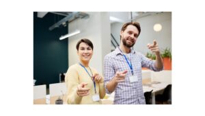 Two professionals wearing ID badges in a modern office, pointing towards the camera in a welcoming manner